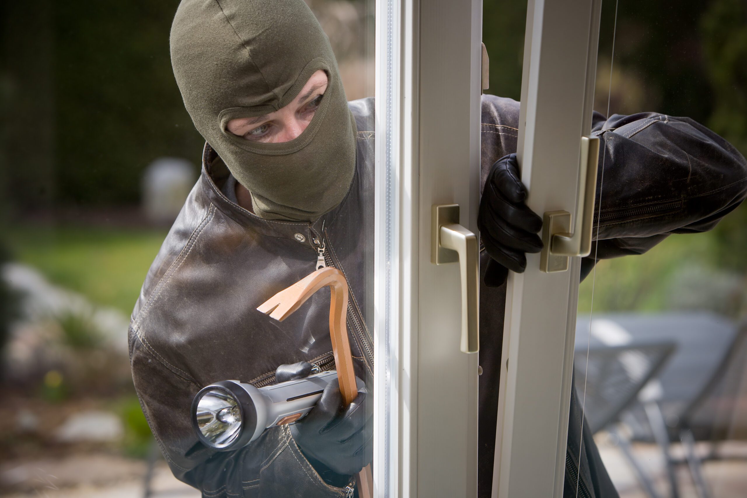 Masked burglar breaking into a home with a crowbar and flashlight, representing the rising trend of home burglaries targeting professional athletes.